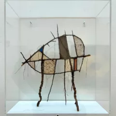 A sculptural object made of twigs, strings and colored papers stands in a glass vitrine, almost like an insect or small creature set for study in a natural history museum.