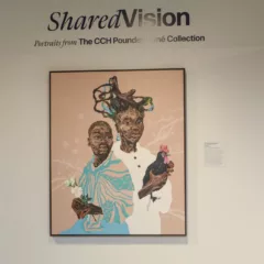 A museum wall shows a multicolored painting framed in black under a sign saying, “Shared Vision: Portraits from The CCH Pounder Koné collection.” The painting shows two Black figures.