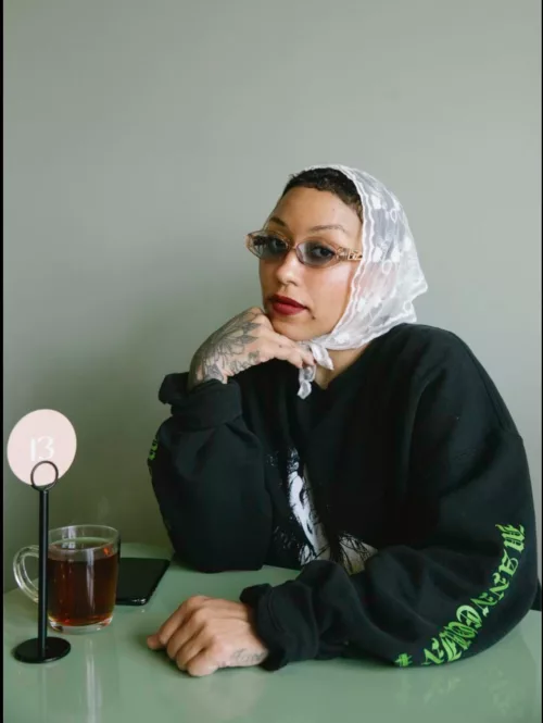 A woman sits at a green formica table in front of a green tinted wall, looking at you with a side eye, wearing dark tinted eyeglasses, a white lace headscarf, red lipstick, black sweatshirt and holding one tattooed hand under her chin, and the other clenched in a fist resting on the table.