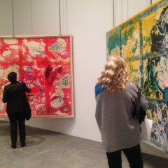 Two large colorful paintings are hung in an art gallery. A woman is standing in front of each work looking at it closely.